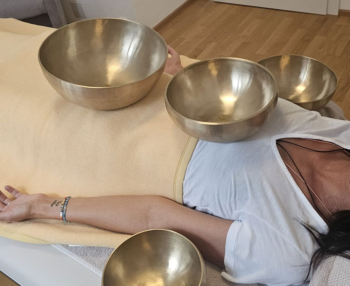 Vera Volkmann - Physiotherapie - Eine Person liegt entspannt auf einer Liege in einem hellen Praxisraum mit Holzboden. Sie trägt ein weißes T-Shirt und ist teilweise mit einer gelben Decke zugedeckt. Drei metallene, goldfarbene Klangschalen sind auf ihrem Körper (Unterleib, Bauch, Brust) platziert. Eine weitere Klangschale liegt neben dem entspannten Arm der Person. Das Bild zeigt eine Klangschalentherapie-Behandlung.